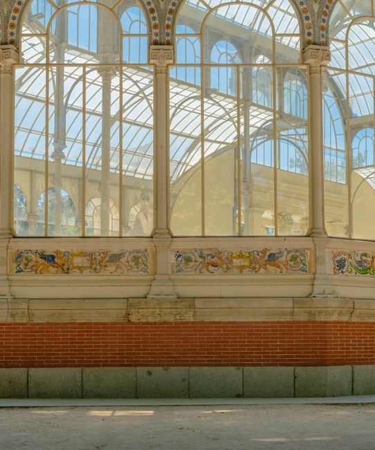 A large glass conservatory with arched window panes and intricate metal framing, illuminated by natural daylight. The interior features a decorative panel with colorful painted motifs along the lower 