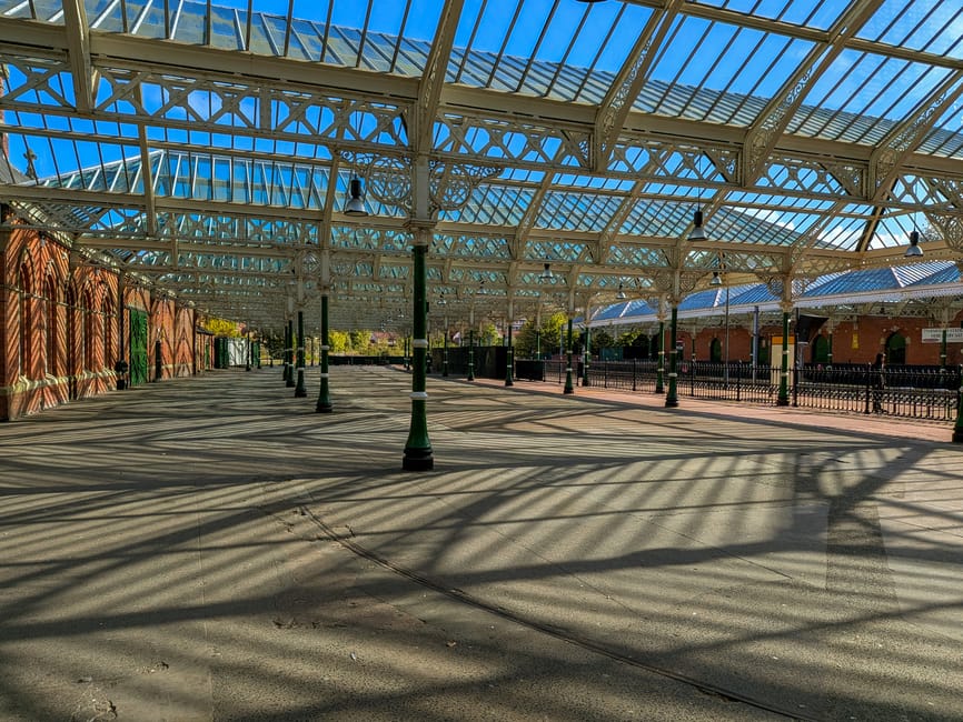 A large glass conservatory with arched window panes and intricate metal framing, illuminated by natural daylight. The interior features a decorative panel with colorful painted motifs along the lower 