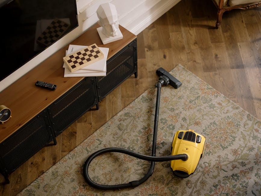 A yellow canister vacuum cleaner with a black hose and extension wand is positioned on a patterned area rug in a living room. To the left, a wooden console table with black metal legs holds a white bu