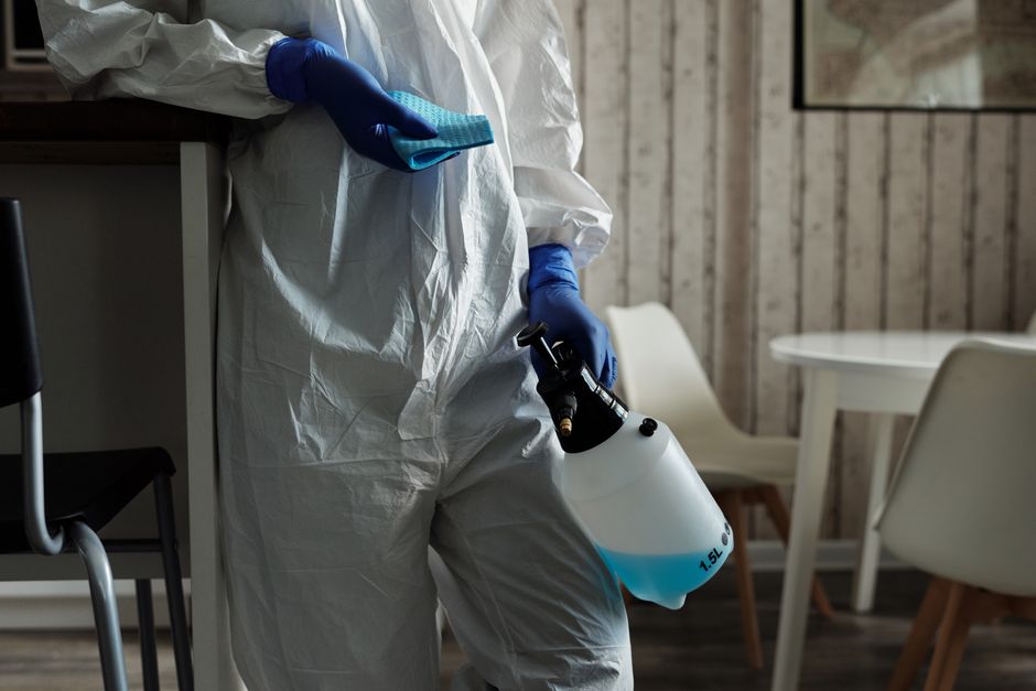 A professional cleaner wearing a grey t-shirt and red uniform apron disinfects a stainless steel surface in an indoor setting, using a handheld spray device. The cleaner holds a blue bucket with a han